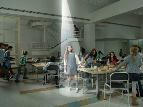 A young girl stands under a beam of light in a school cafeteria, surrounded by other students and tables, creating a surreal scene.