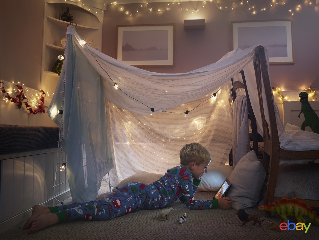 Young boy in pajamas lying on the floor inside a blanket fort, illuminated by string lights, looking at a tablet in a cozy room.