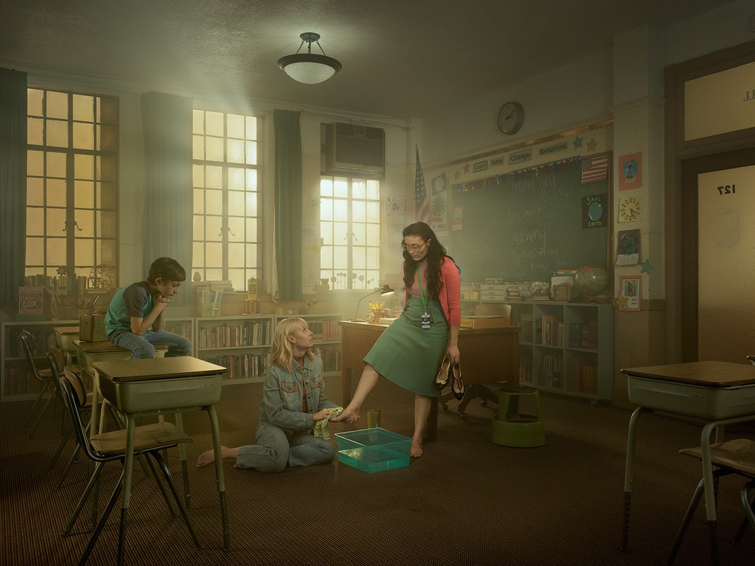 Inside a classroom, a teacher stands barefoot near a small tub of water while two kids, a boy and a girl, sit nearby, with a chalkboard.
