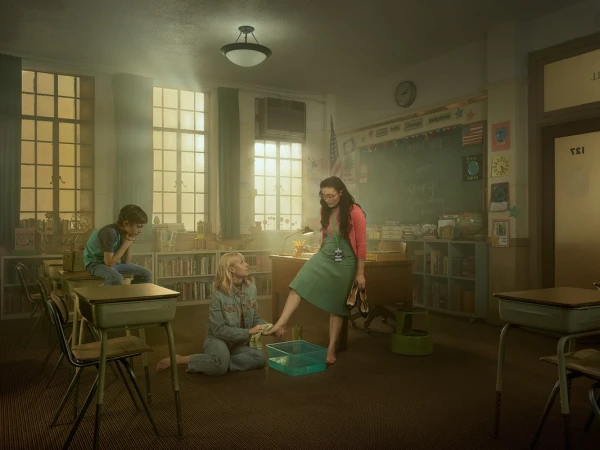 Inside a classroom, a teacher stands barefoot near a small tub of water while two kids, a boy and a girl, sit nearby, with a chalkboard.