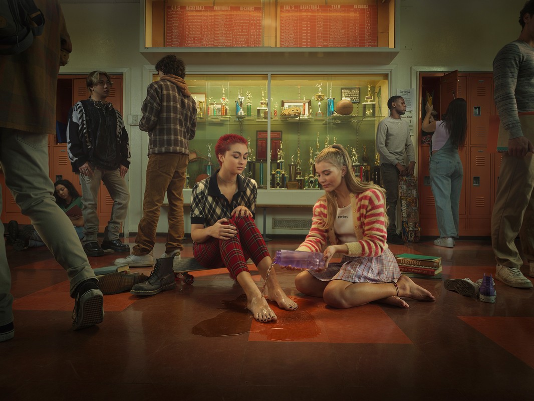 Two young women sit on the floor in a school hallway surrounded by students. A woman pours liquid on the other's foot.