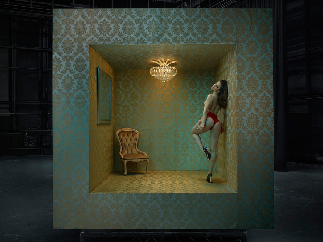 Nude woman in red lingerie leans against the floral wallpaper inside a framed cube, illuminated by a chandelier in the interior space.