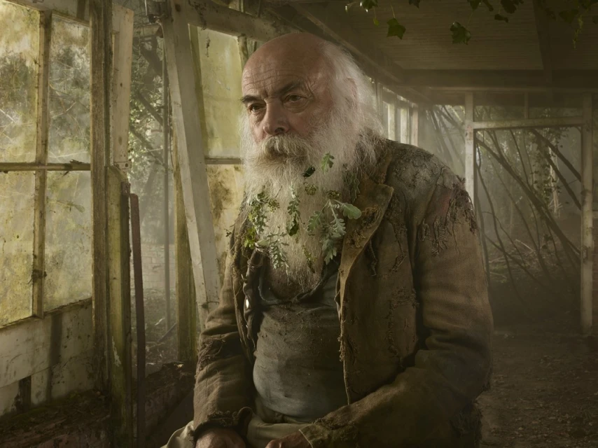 Elderly man with a long white beard wearing tattered clothing and leaves, standing inside a greenhouse, portrait photograph.