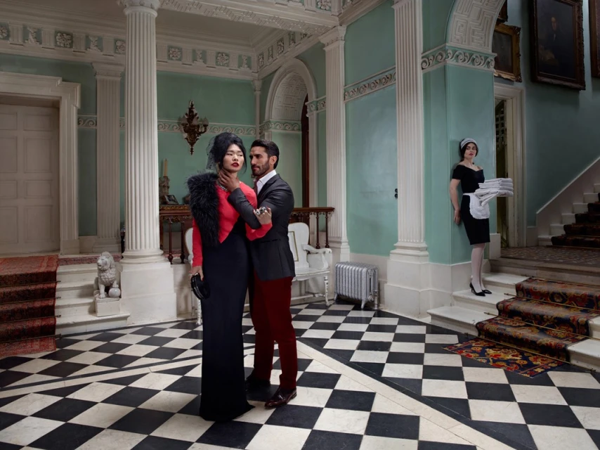 Elegant interracial couple embraces inside a grand mansion hall with a maid in the background, showcasing a scene of luxury and classic style.