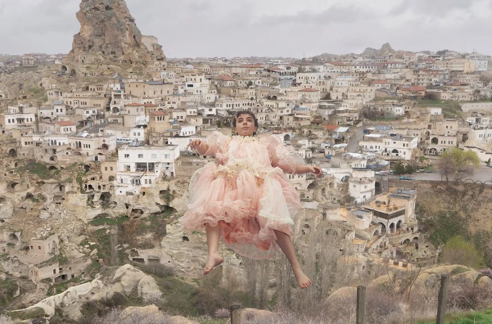 Young woman in a pink dress seemingly floats mid-air against the backdrop of Cappadocia's unique geological formations and dwellings.