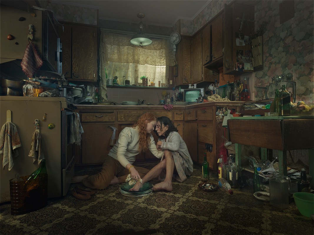 Two women seated on a carpeted kitchen floor, one gingerly tending to the other's injured foot, surrounded by various household objects.