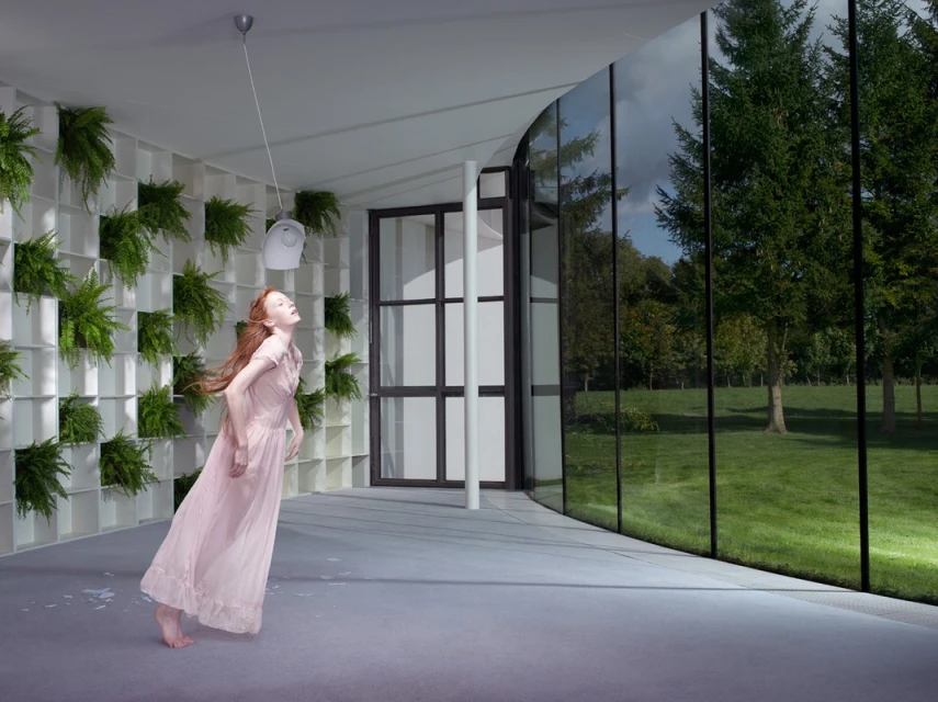 Woman in pink dress poses in minimalist architecture space with fern wall, open black framed doors and windows looking to trees.