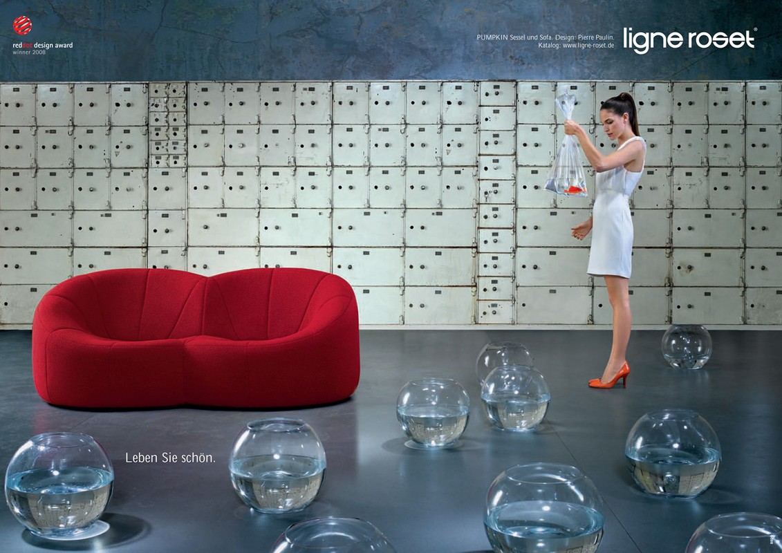 Interior design advertisement: a woman in a white dress holding transparent bags stands near a red sofa and clear glass bowl vases.