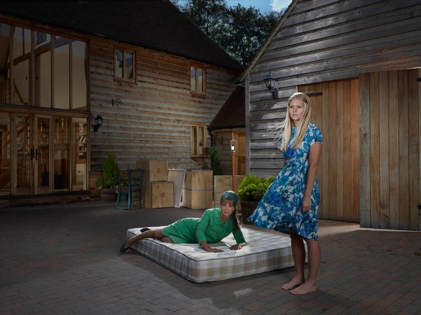 Two women in dresses pose in an outdoor setting beside a mattress on a brick surface, with wooden buildings in the background.
