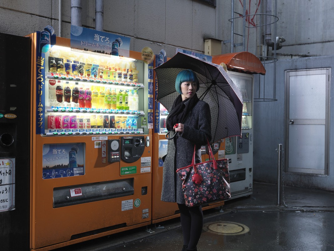 Woman with blue hair stands next to vending machines in an urban setting, holding an umbrella and a floral handbag on a wet day.