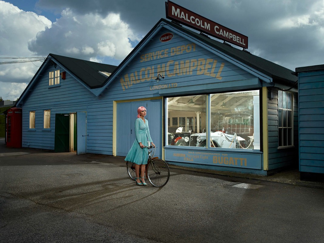 Vintage-style image of a woman with pink hair on a bicycle in front of a blue building with 'Malcolm Campbell' sign. Cars inside the building.