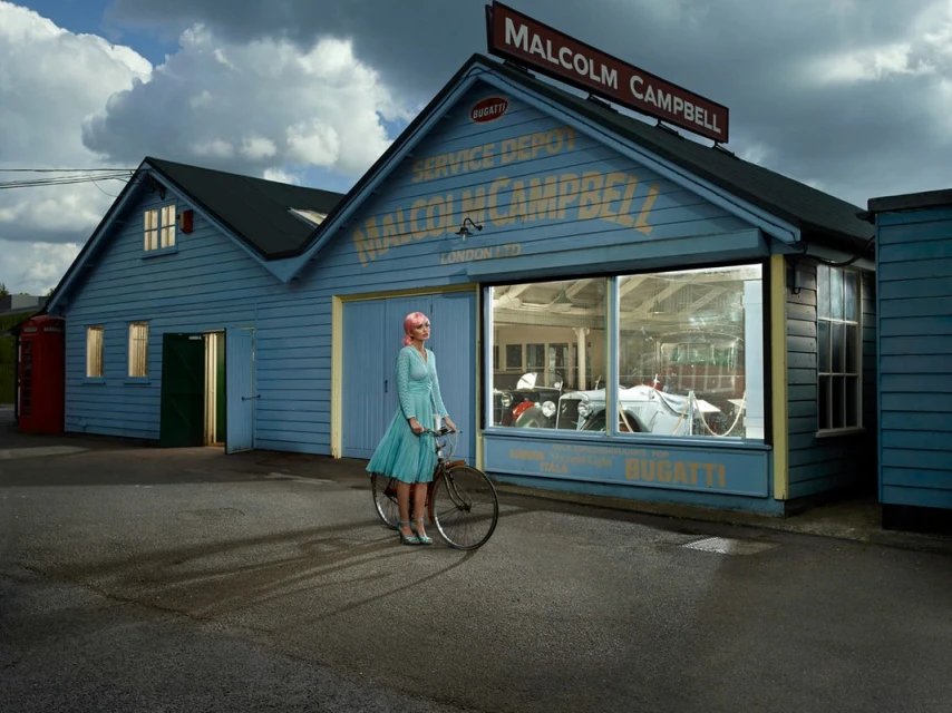 Vintage-style image of a woman with pink hair on a bicycle in front of a blue building with 'Malcolm Campbell' sign. Cars inside the building.