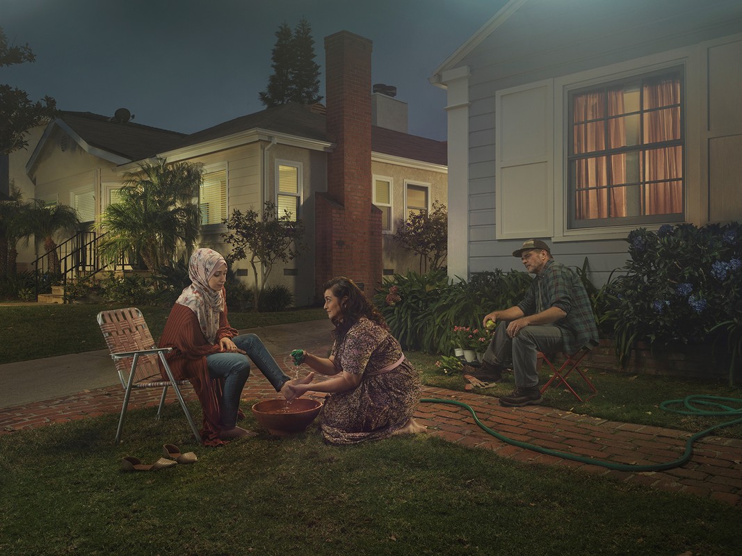 Woman washing another woman's feet in an outdoor setting near a house, with a man seated nearby, creating a family-centered scene.
