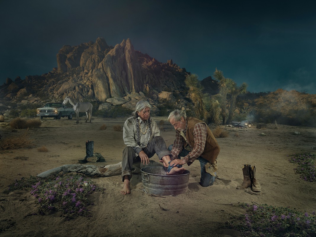 Older man washing the feet of an older woman in a metal tub, outdoors in the desert with a vehicle and rock formations in background.