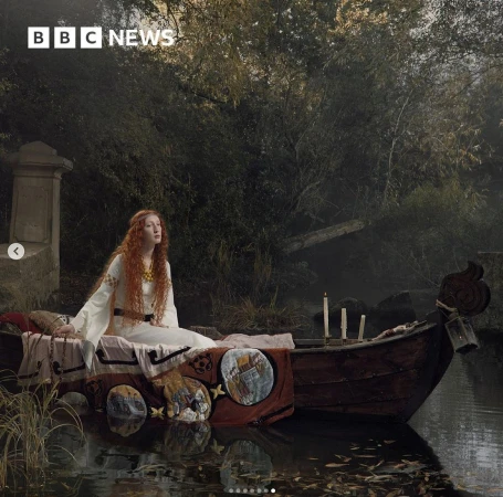 Red-haired woman in a boat on a pond in front of trees. She is sitting up and looking up, with candles on the boat.