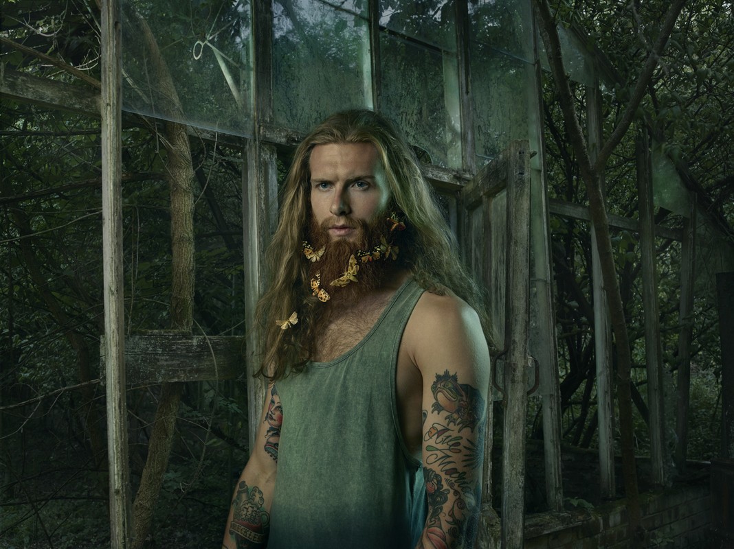 Portrait of adult male with long brown hair and beard decorated with butterflies, wearing a tank top in an overgrown greenhouse.