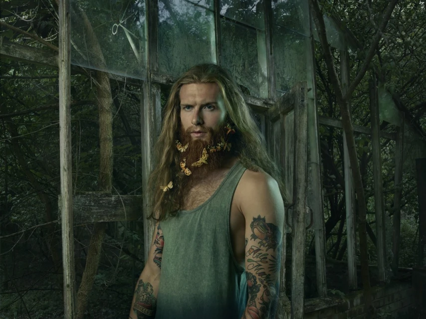Portrait of adult male with long brown hair and beard decorated with butterflies, wearing a tank top in an overgrown greenhouse.