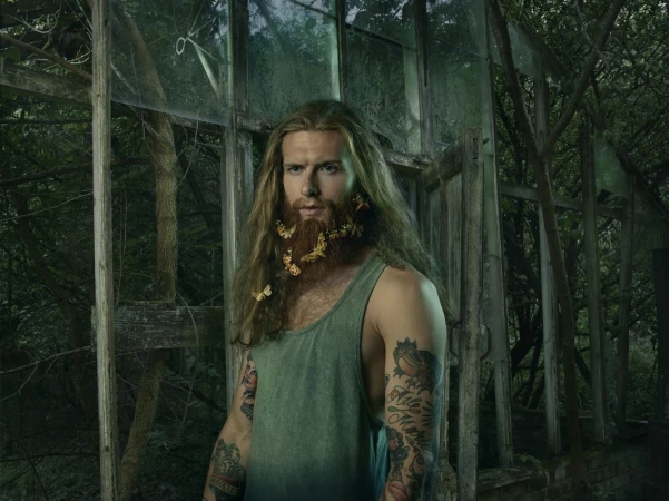 Portrait of adult male with long brown hair and beard decorated with butterflies, wearing a tank top in an overgrown greenhouse.