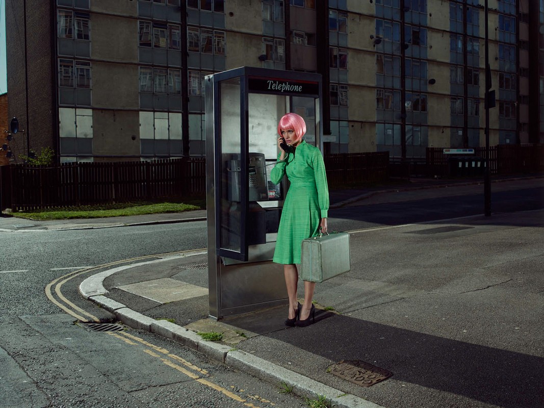 Stylish woman in green dress and pink wig uses phone booth on urban corner, carrying briefcase against a backdrop of buildings.