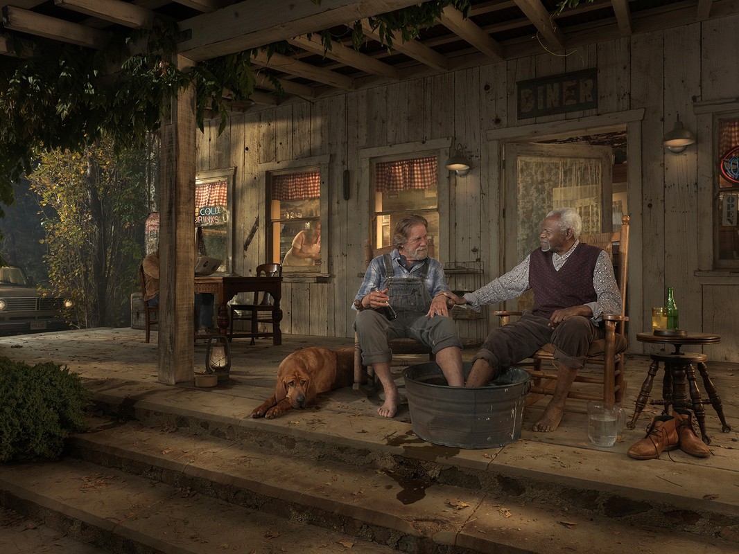 Two older men sit on a porch, one soaks his feet in a tub, accompanied by a dog, with a vintage car and building in the background.