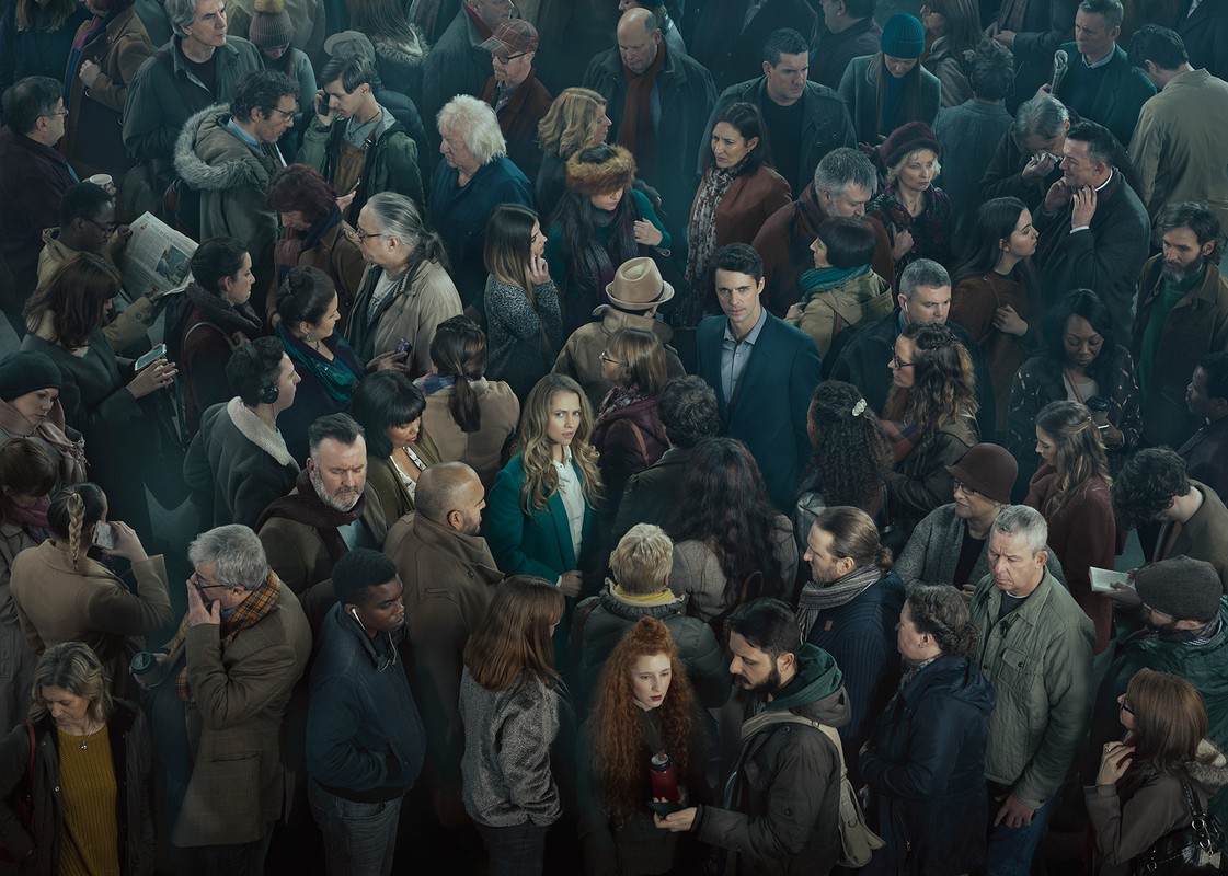 Diverse crowd of men and women, close together, filling the frame with an emphasis on multiple faces and varied clothing styles.