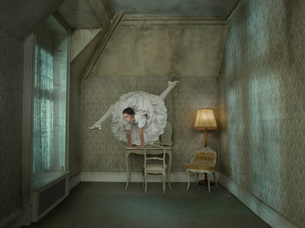 A young woman in a white dress strikes a handstand-like pose on a table in an ornate room with patterned wallpaper, bathed in diffused light.