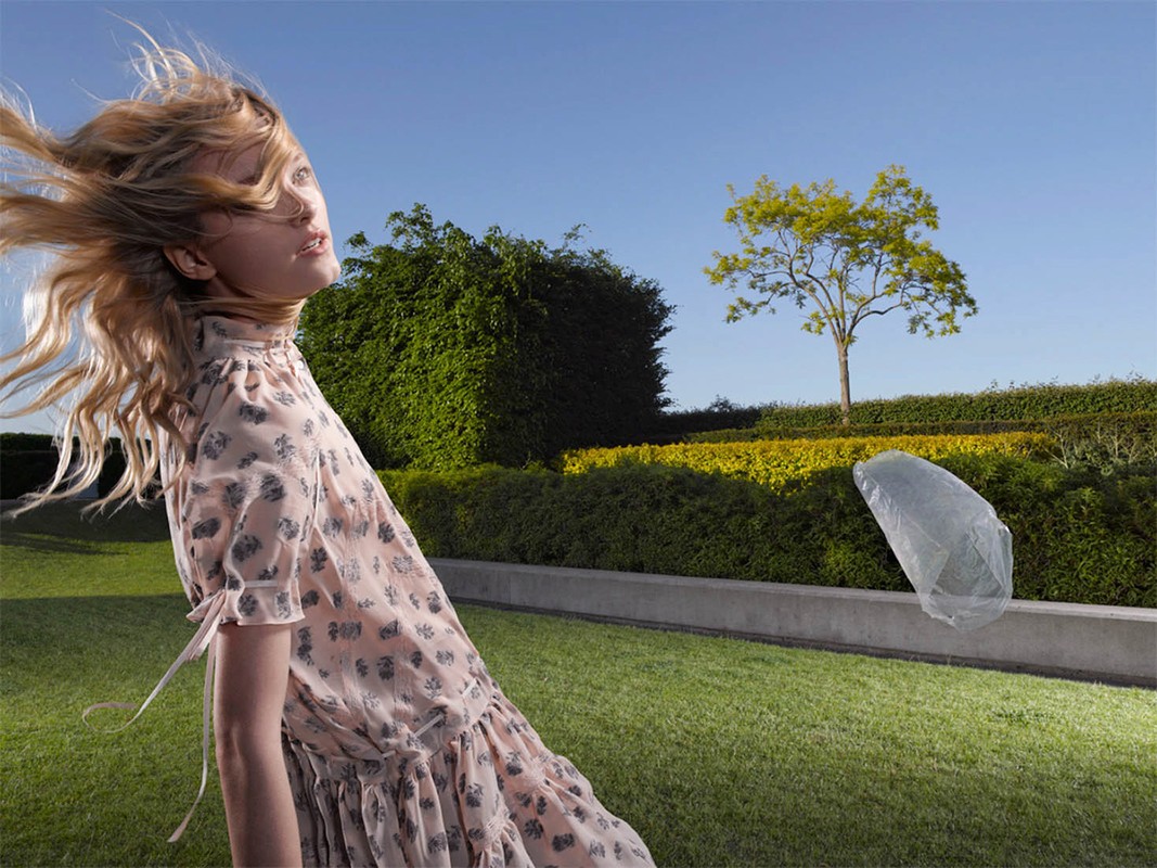 Blonde fashion model with flowing hair wearing a patterned dress poses outdoors in a garden on a sunny day, with greenery in the background.