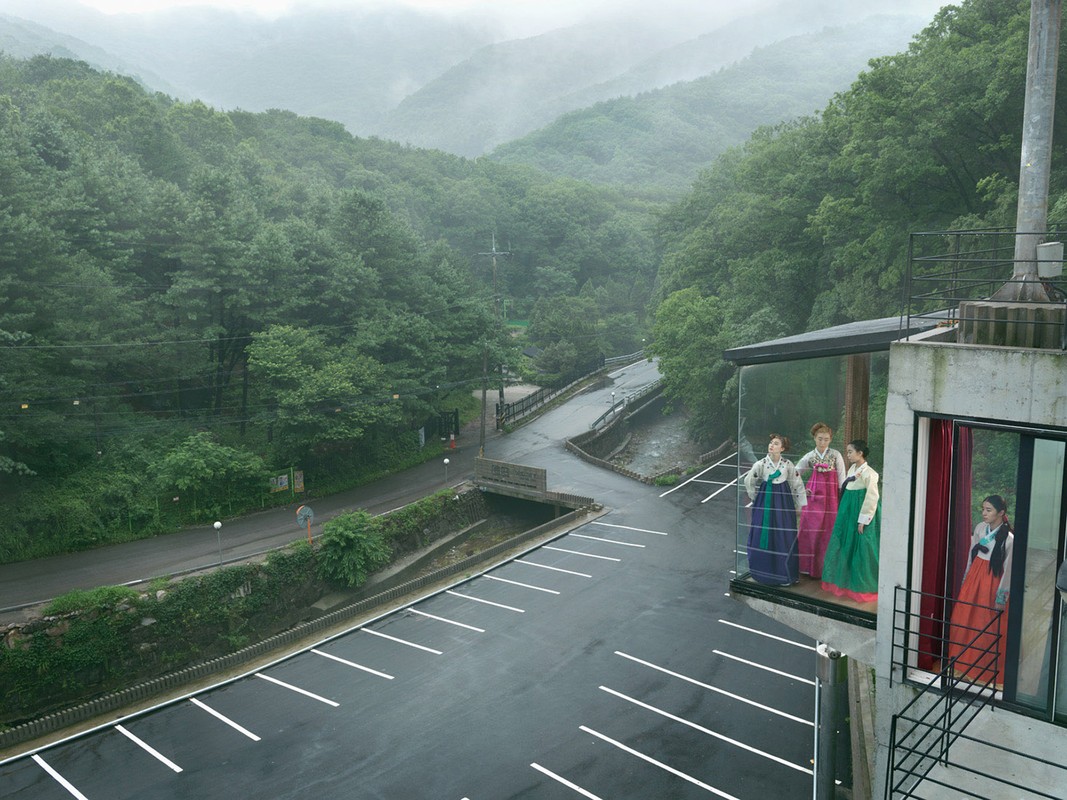 Group of women wearing colorful hanboks standing on a building's balcony with mountains and greenery in the distance under an overcast sky.