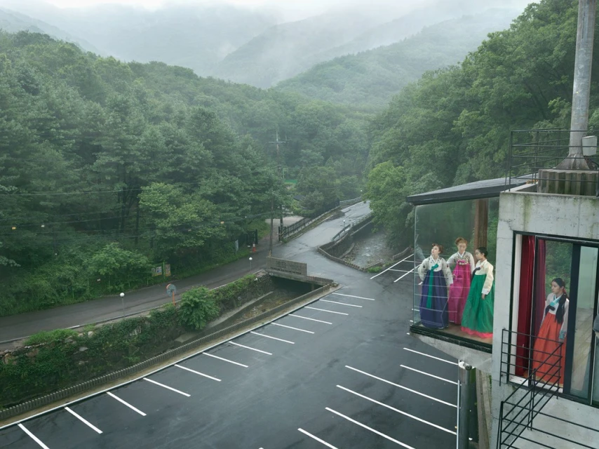Group of women wearing colorful hanboks standing on a building's balcony with mountains and greenery in the distance under an overcast sky.