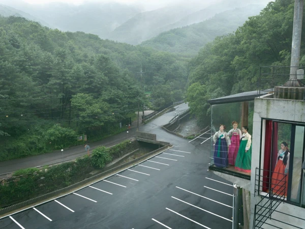Group of women wearing colorful hanboks standing on a building's balcony with mountains and greenery in the distance under an overcast sky.