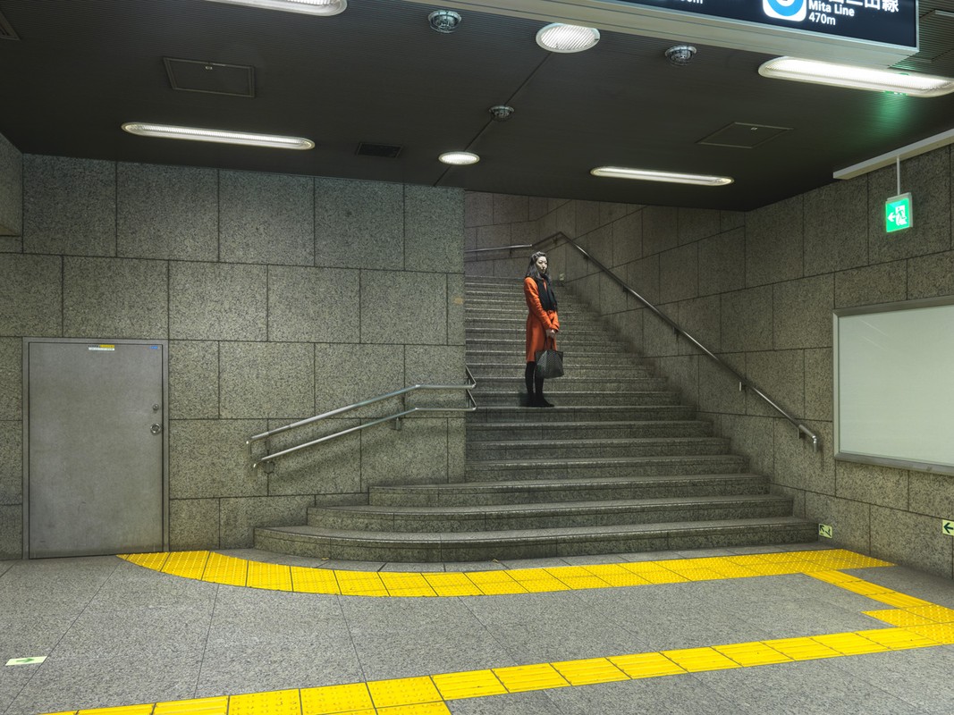 Woman in orange coat ascends subway stairs with yellow safety markings. Interior public transit station with grey tiled walls and lighting.