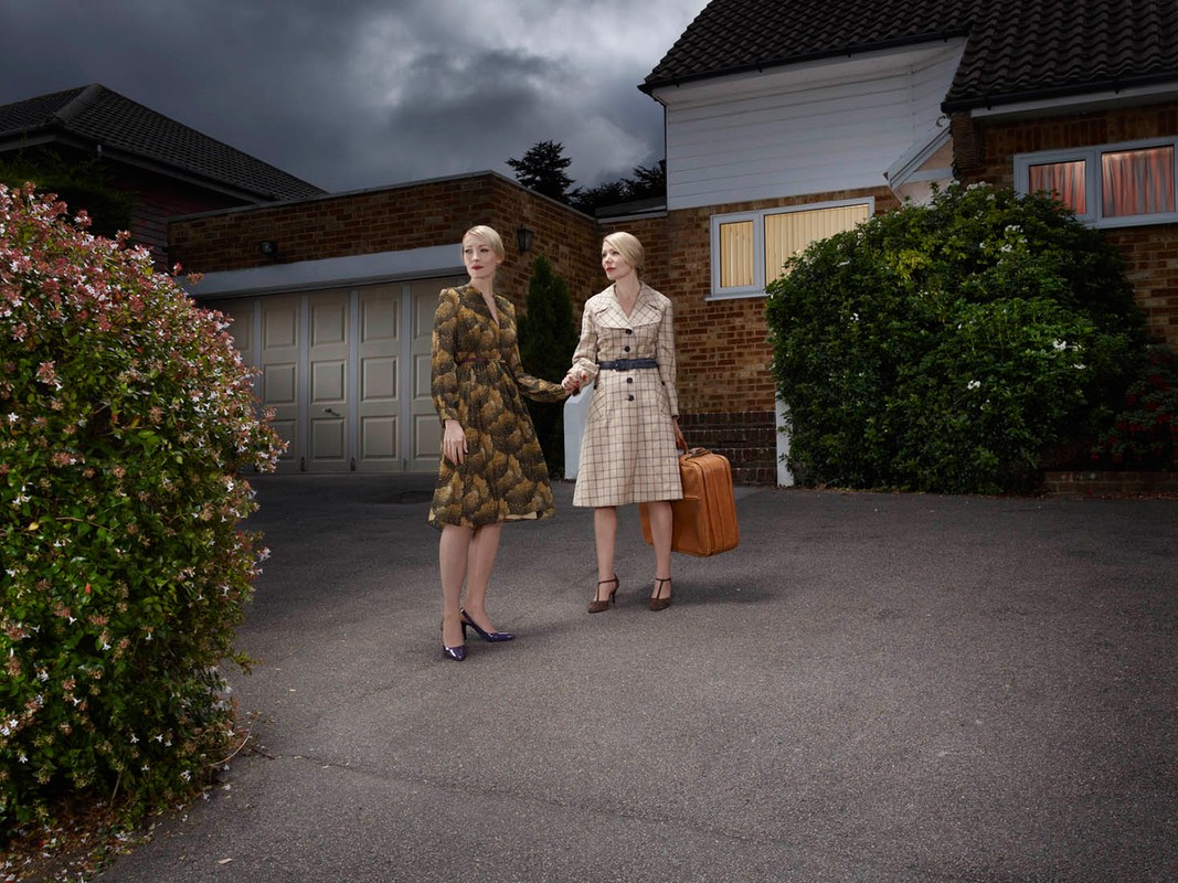 Two adult women in vintage-style dresses standing in front of a house with a suitcase on a dark, cloudy day in an outdoor setting.