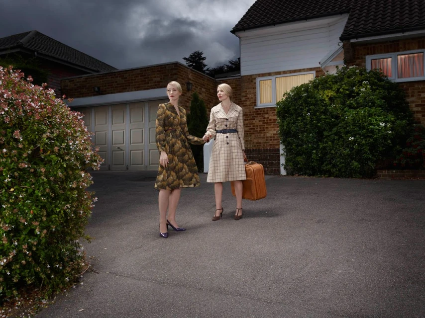 Two adult women in vintage-style dresses standing in front of a house with a suitcase on a dark, cloudy day in an outdoor setting.