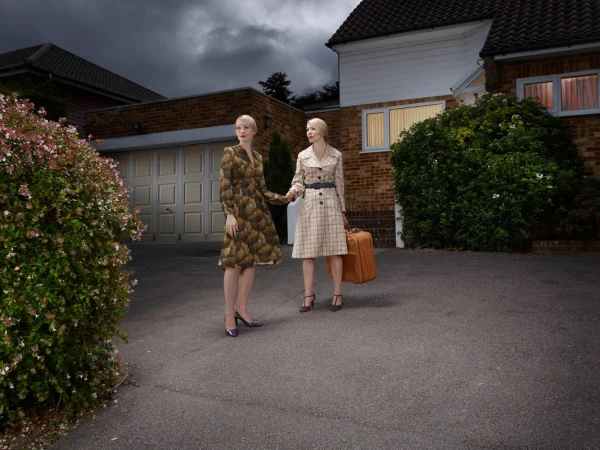 Two adult women in vintage-style dresses standing in front of a house with a suitcase on a dark, cloudy day in an outdoor setting.