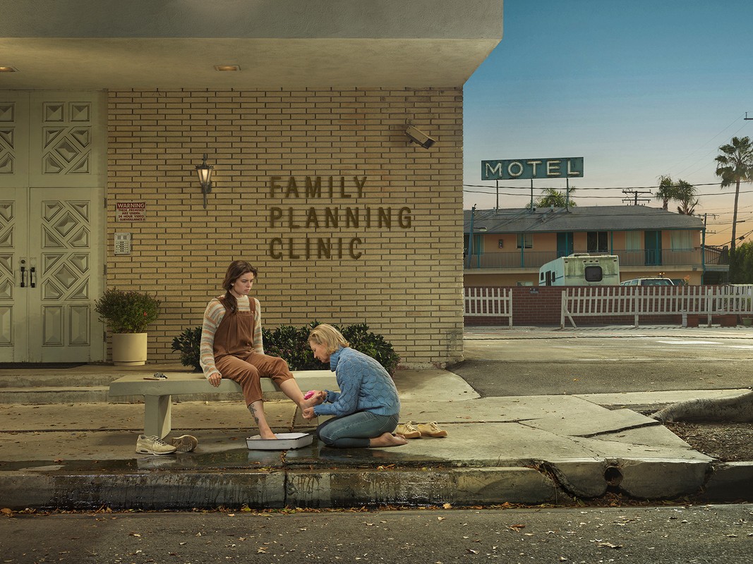 Young woman getting her foot washed by an adult on a bench outside of a clinic in front of a motel against a blue sky.