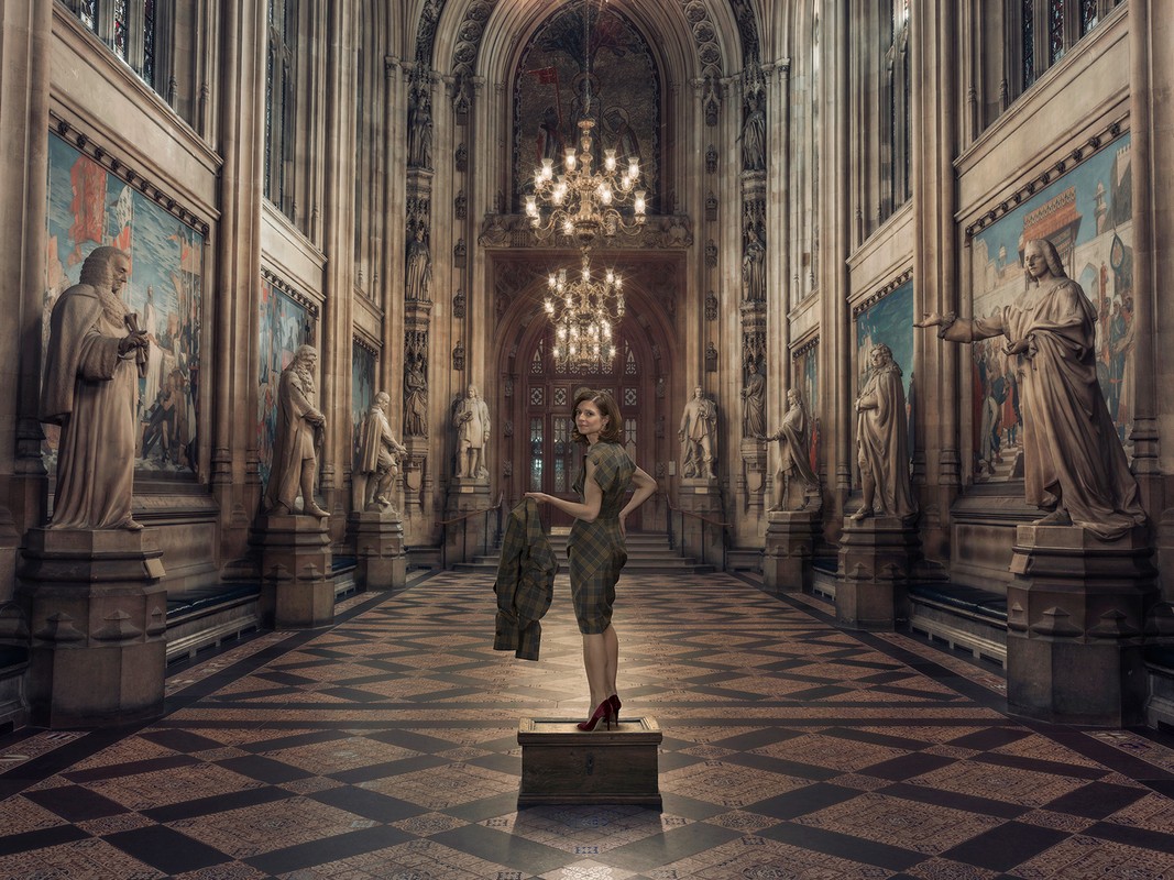 An adult woman stands on a box in a grand hall with statues, paintings, and chandeliers, holding a coat and wearing red heels.