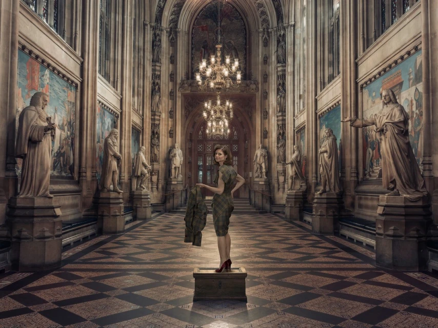 An adult woman stands on a box in a grand hall with statues, paintings, and chandeliers, holding a coat and wearing red heels.