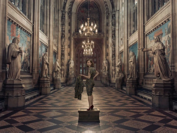 An adult woman stands on a box in a grand hall with statues, paintings, and chandeliers, holding a coat and wearing red heels.
