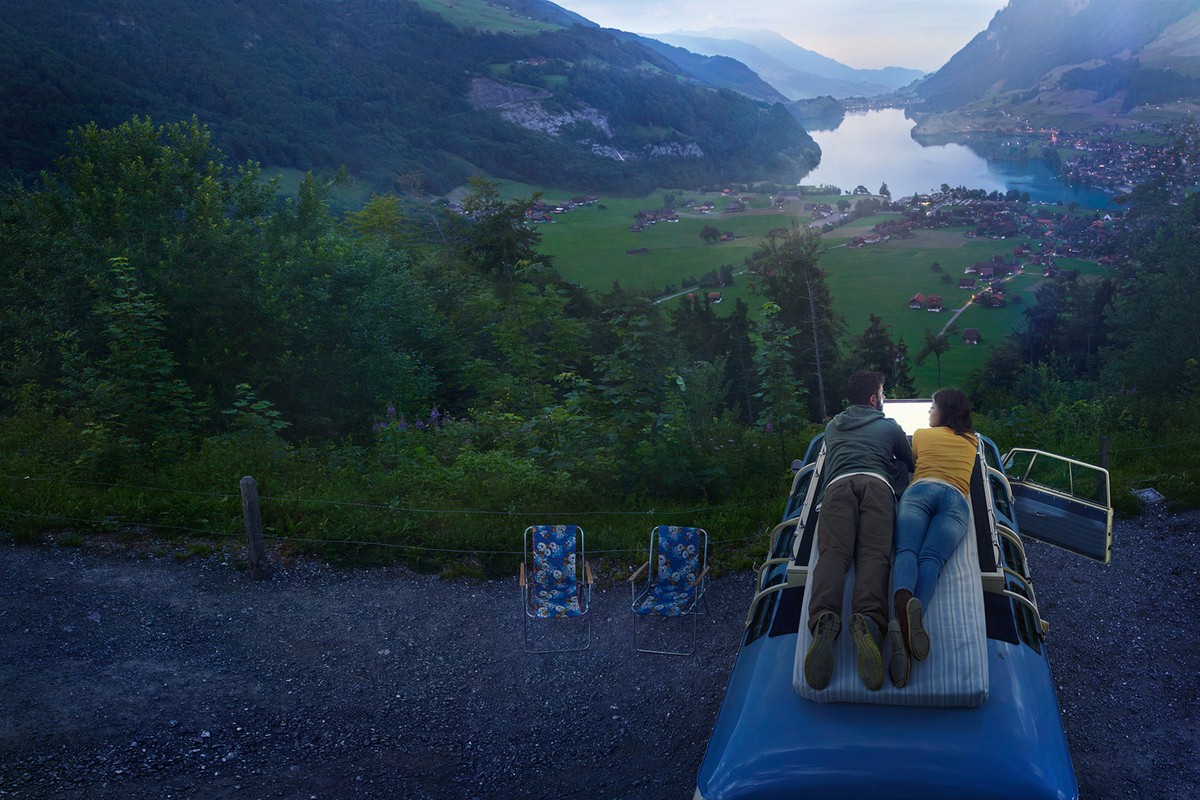 Couple lounging on a van roof with a mountain and lake view, chairs beside the van, creating a scenic and relaxing travel moment.
