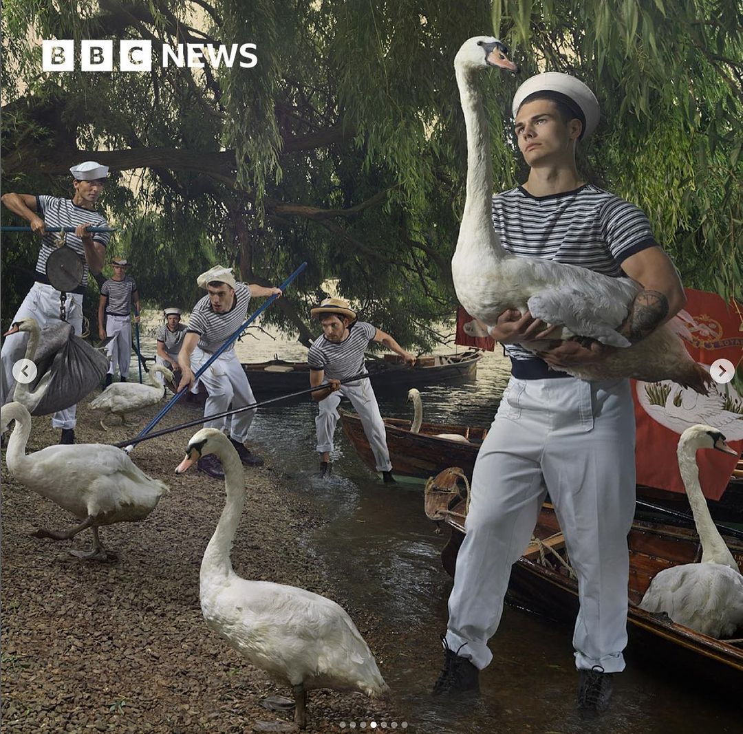 Group of men in sailor outfits with swans by the water, BBC News logo in corner, vintage-style editorial photography.