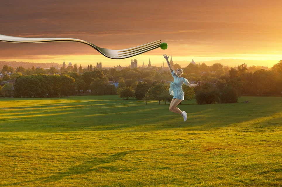 Woman jumping in a field toward giant fork with pea, set against a golden sunset and cityscape backdrop. Food and fun concept.