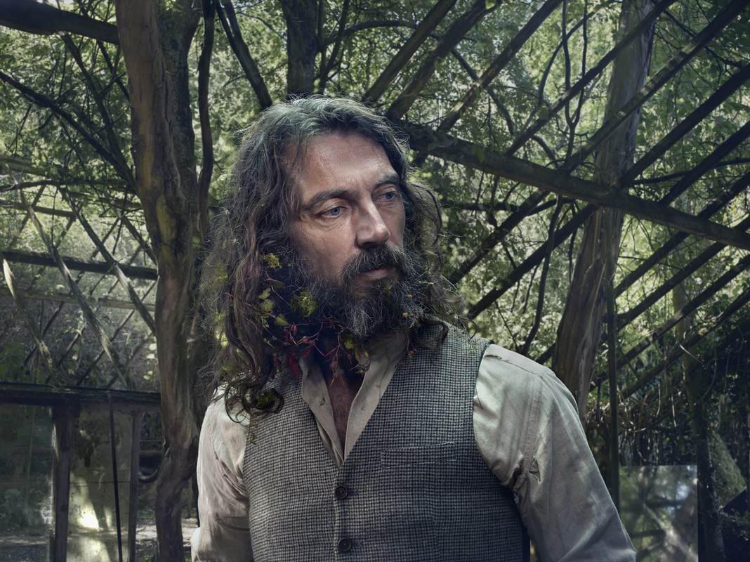 Portrait of an adult male with long hair and a beard with vines in a rustic vest, set against a backdrop of trees and greenhouse.