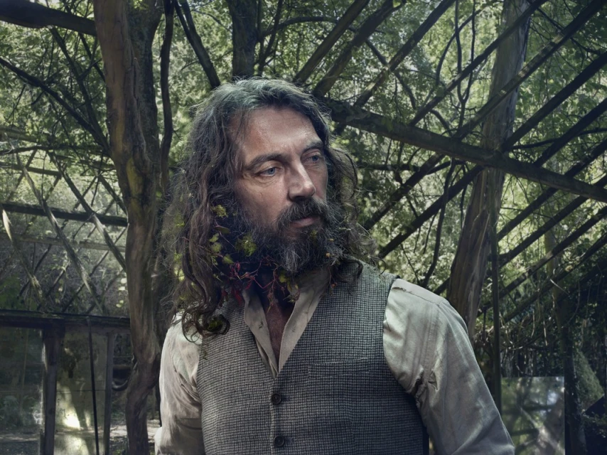 Portrait of an adult male with long hair and a beard with vines in a rustic vest, set against a backdrop of trees and greenhouse.
