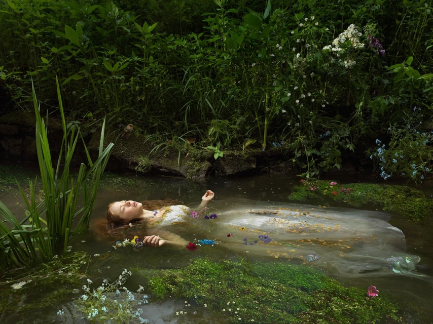 A long-haired woman wearing an off-white dress floats in a stream with greenery and flowers surrounding her, creating a serene scene.