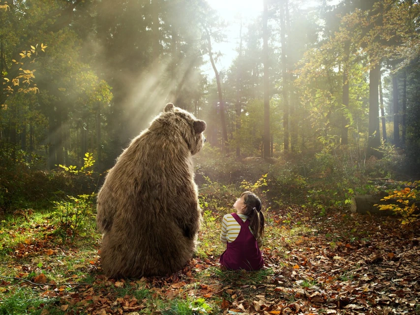 Back view of a girl sitting cross-legged in a forest, looking up at a standing brown bear in sunlight, creating a whimsical scene.
