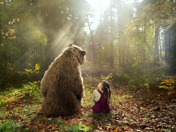 Back view of a girl sitting cross-legged in a forest, looking up at a standing brown bear in sunlight, creating a whimsical scene.