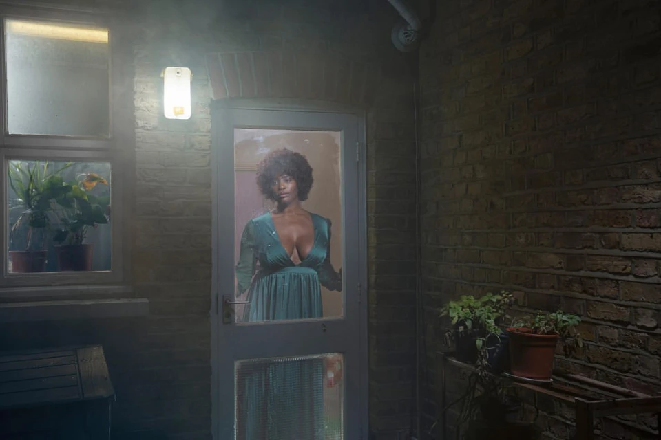 A young Black woman with an afro stands in a doorway wearing a green dress, plants and brick visible around her in soft lighting.