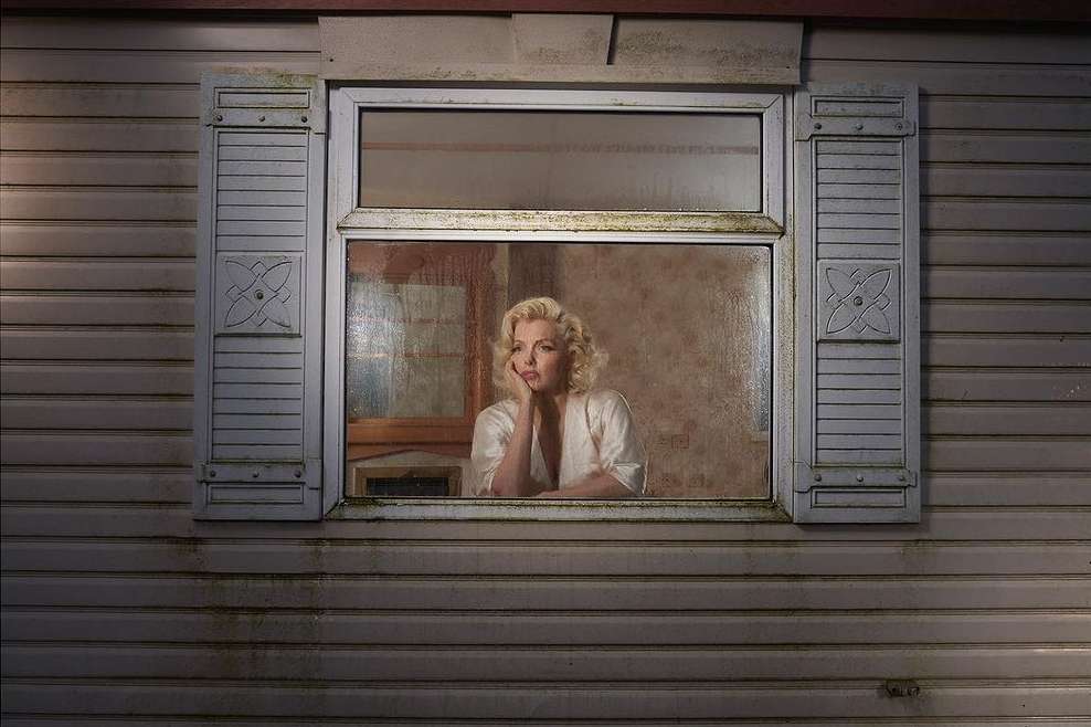 Adult female with blonde hair gazes out of a grimy window in an exterior shot of a grey wooden home with light from inside.