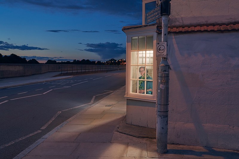 Adult female stands inside brightly lit room with window to sidewalk at dusk, street and treeline visible in background, streetlight in view.
