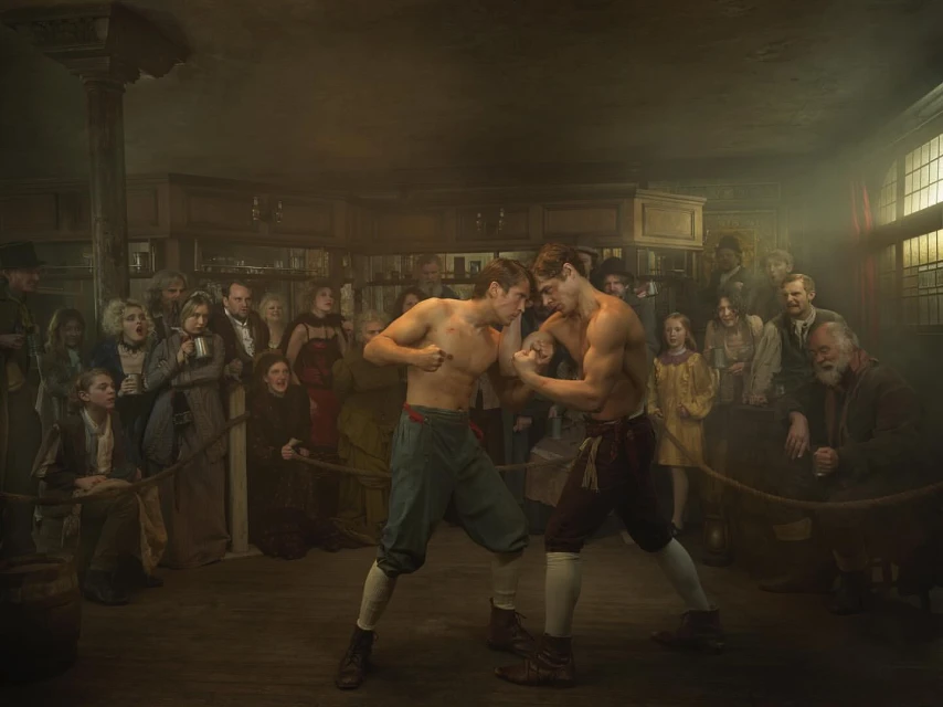 Two shirtless male boxers face off in an indoor ring surrounded by a crowd in vintage clothing. The scene is dimly lit, creating a dramatic effect.